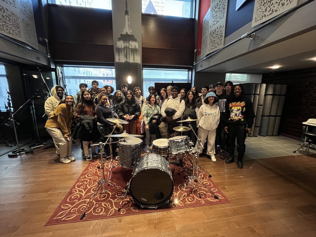 Spring 2023 Future Music Moguls students pose for a group photo, standing behind a drum kit in a recording studio